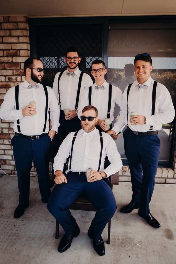 The groom and four groomsmen dressed in white shirts, navy pants, gray bow ties, and black suspenders pose together outside Ocean View Estates, each holding a canned drink. The groom is seated in a chair wearing sunglasses, while the groomsmen stand behind him.