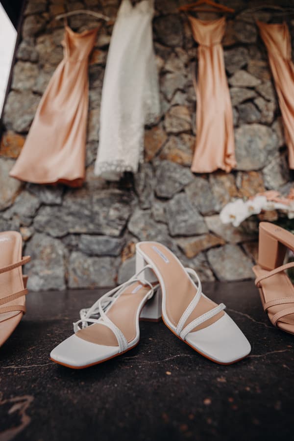 Bride's white heeled shoes placed on a dark surface with peach bridesmaid dresses and a white wedding dress hanging against a stone wall at Preston Peak Winery Functions Centre.
