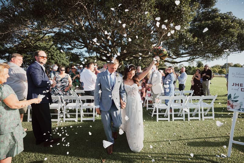 Bride Jheng and groom Dane walk down the aisle at Sandstone Point Hotel — The Pavilion as guests throw white flower petals.