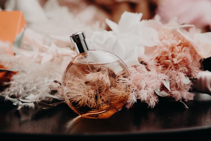 A bottle of Hermès perfume placed on a dark surface with a soft-focus background of pink and white floral arrangements at Royal on the Park.