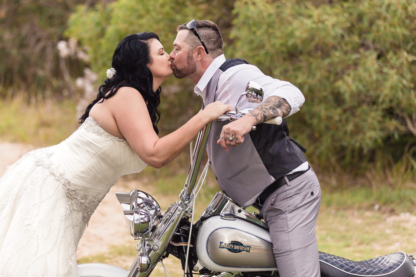 The bride Kelly and groom Shane kiss while Shane sits on a Harley-Davidson motorcycle outdoors. Destination wedding photographer by She Said Yes.