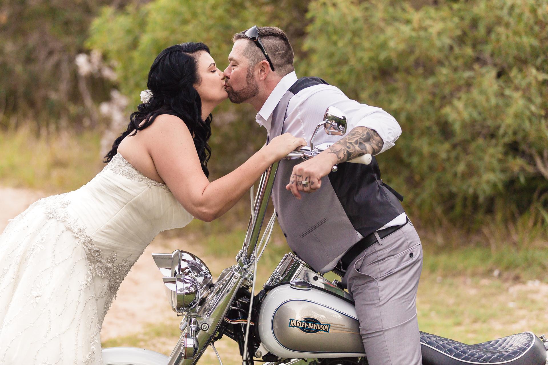 The bride Kelly and groom Shane kiss while Shane sits on a Harley-Davidson motorcycle outdoors at Novotel Sunshine Coast Resort.
