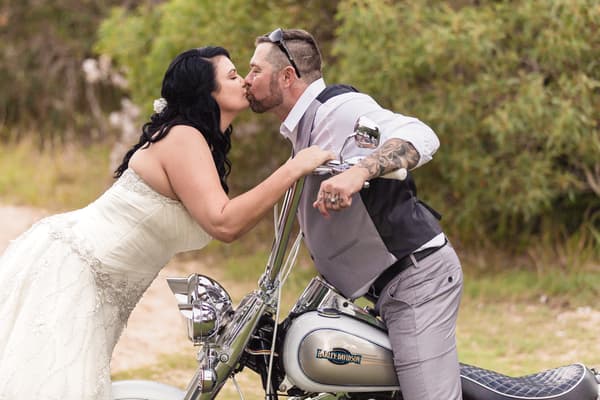 The bride Kelly and groom Shane kiss while Shane sits on a Harley-Davidson motorcycle outdoors. Best time of year to get married by She Said Yes.