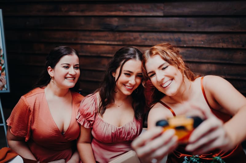 Three female guests at the reception stage of, smiling and taking a selfie together in front of a wooden wall. What is an engagement party by She Said Yes.
