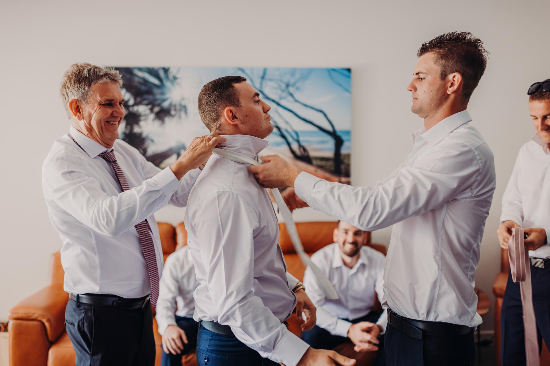 The groom is being helped with his collar and tie by the father of the groom and a groomsman in a preparation room with other men seated in the background.