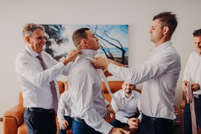 The groom is being helped with his collar and tie by the father of the groom and a groomsman in a preparation room with other men seated in the background.