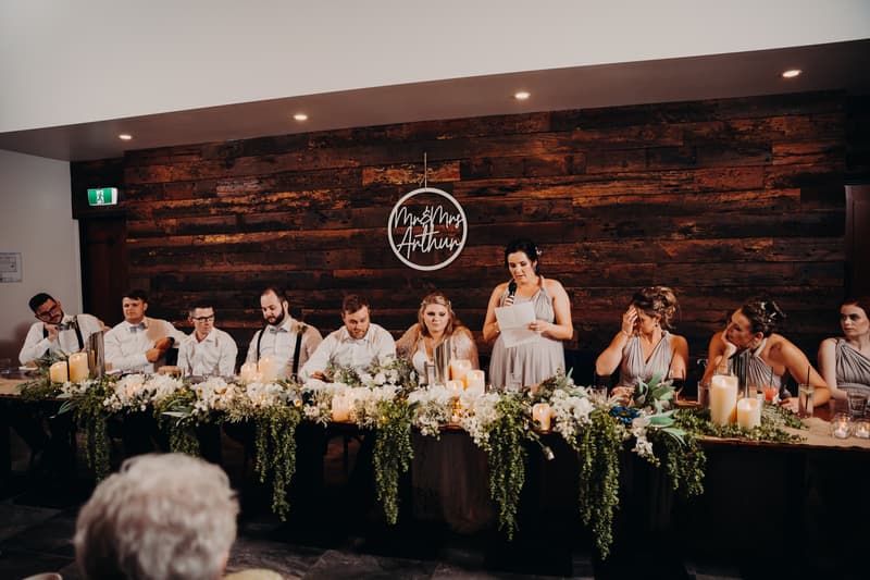Bridal party seated at a long table decorated with white flowers and candles at Ocean View Estates, with a bridesmaid standing and reading a speech.