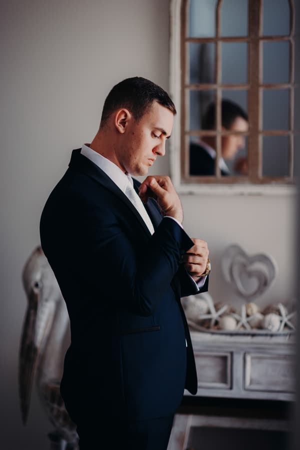 The groom Dylan adjusts his suit jacket in front of a mirror inside a room at Sandstone Point Hotel.