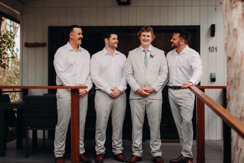 The groom and three groomsmen stand together on a wooden deck outside a building numbered 101 at Sandstone Point Hotel.