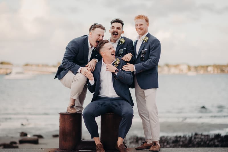 Four groomsmen in navy blazers and light-colored pants pose playfully near water at Sandstone Point Hotel.