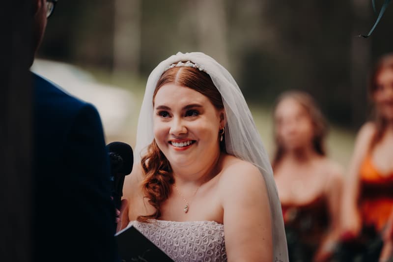 The bride Lilly smiles while holding a microphone during the wedding ceremony at Yabbaloumba Retreat — By The River, with the groom Connor partially visible and bridesmaids blurred in the background.
