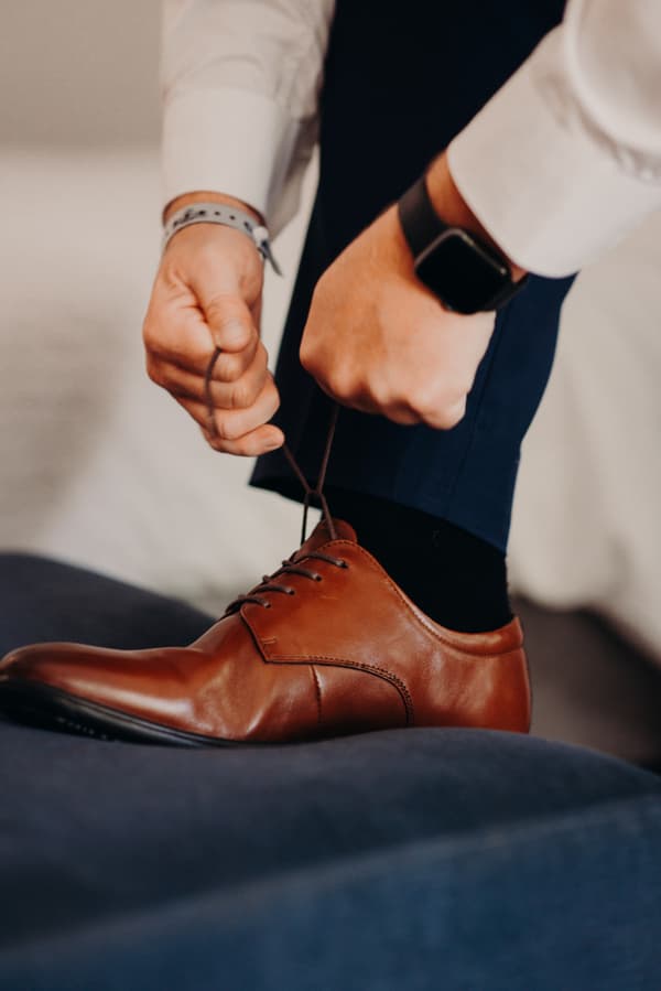 The groom ties the laces of his brown leather dress shoe while seated, wearing dark blue trousers and a white shirt.