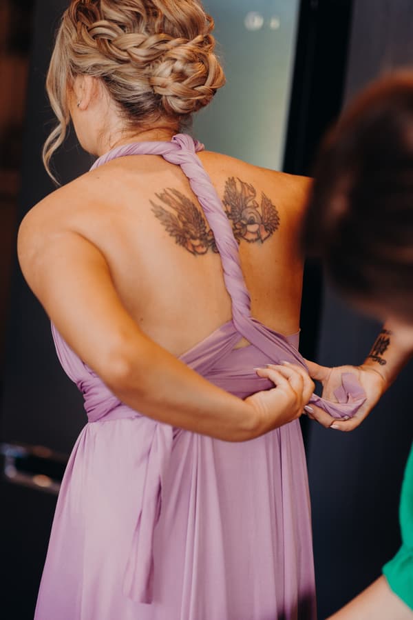 The bride is having the back of her lavender dress adjusted by another person indoors.