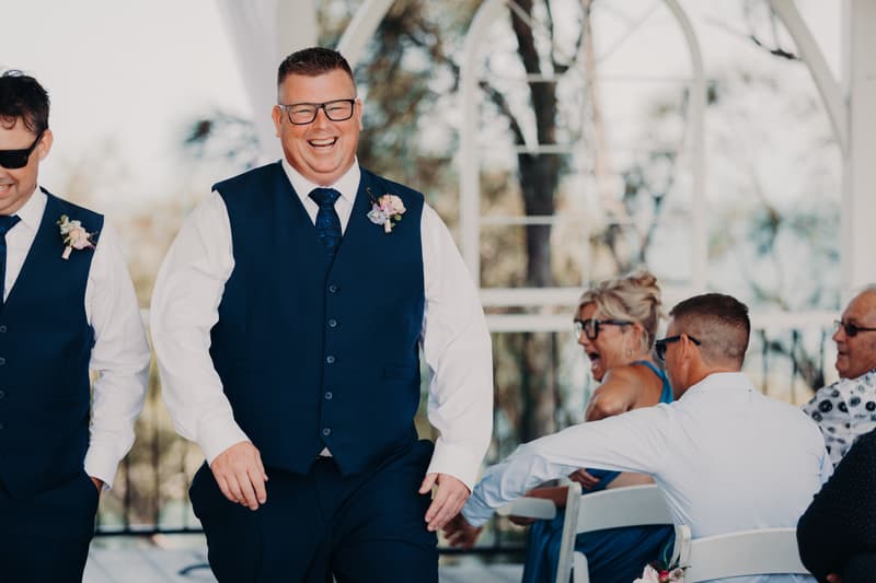 Peter, the groom, wearing a navy vest and tie with a boutonniere, smiles broadly while walking at the Sandstone Point Hotel — Pavilion during the ceremony. A groomsman in similar attire is partially visible to his left. Seated guests are visible in the background.
