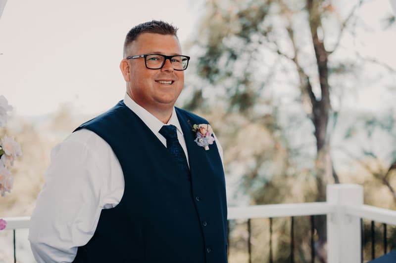 Peter, the groom, wearing a navy vest, white shirt, and tie with a boutonniere, stands smiling at the Sandstone Point Hotel — Pavilion during the wedding ceremony.