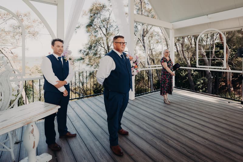 The groom and a groomsman stand on the pavilion stage at Sandstone Point Hotel, with a female celebrant holding a book nearby, surrounded by floral arrangements and overlooking trees.