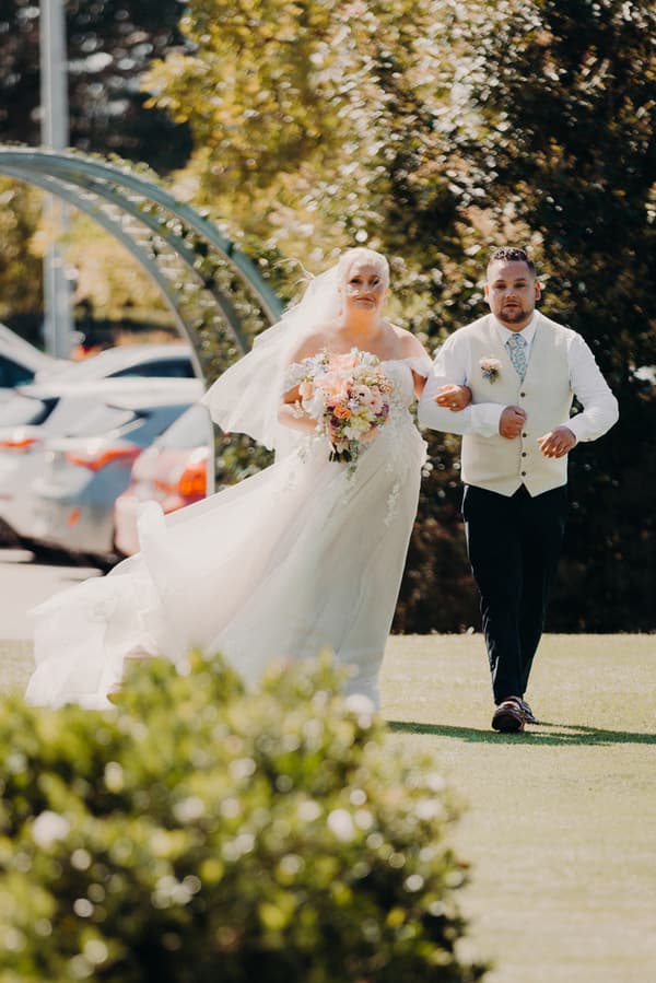The bride Chantelle in a white wedding gown holding a bouquet walks arm-in-arm with the groom Peter, who is wearing a white vest and tie, outdoors at Sandstone Point Hotel — Pavilion.