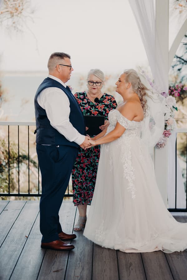 Bride Chantelle and groom Peter hold hands facing each other during their wedding ceremony at Sandstone Point Hotel — Pavilion, with the officiant standing between them reading from a tablet.