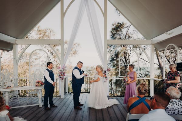 Bride Chantelle and groom Peter stand facing each other at the altar of Sandstone Point Hotel — Pavilion during their wedding ceremony, accompanied by a bridesmaid holding a bouquet, a groomsman, and an officiant reading from a book. Guests are seated watching the ceremony.