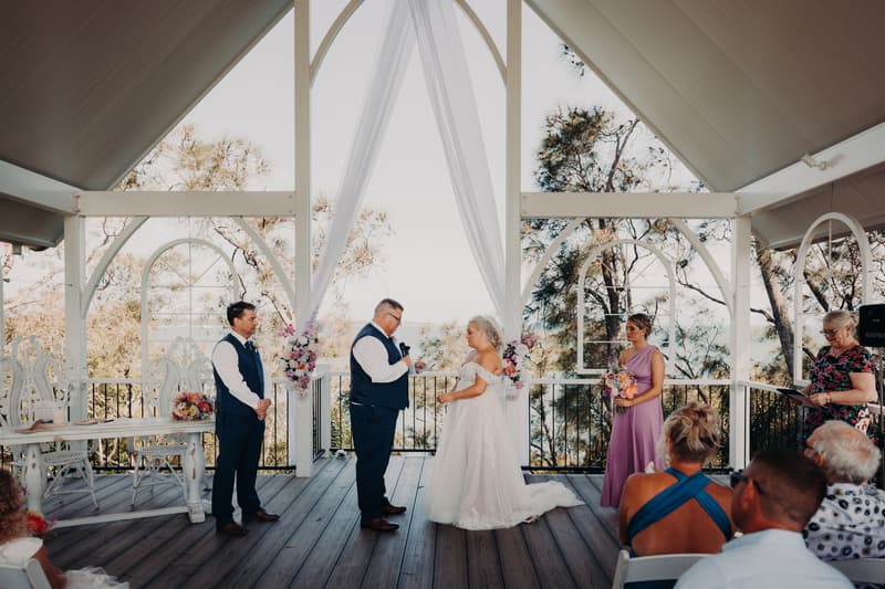 Bride Chantelle and groom Peter stand facing each other at the altar of Sandstone Point Hotel — Pavilion during their wedding ceremony, accompanied by a bridesmaid holding a bouquet, a groomsman, and an officiant reading from a book. Guests are seated watching the ceremony.