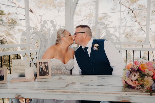 Bride Chantelle and groom Peter share a kiss seated at a white wooden table at Sandstone Point Hotel — Pavilion, with framed photos, a candle, and a floral bouquet on the table.