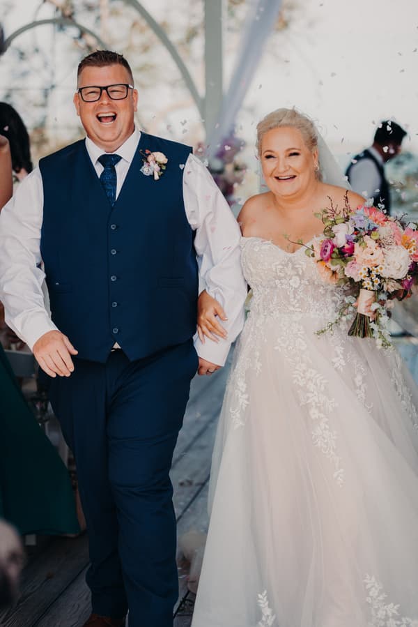 Bride Chantelle and groom Peter walk arm in arm down the aisle at Sandstone Point Hotel — Pavilion, with the bride holding a colorful bouquet.