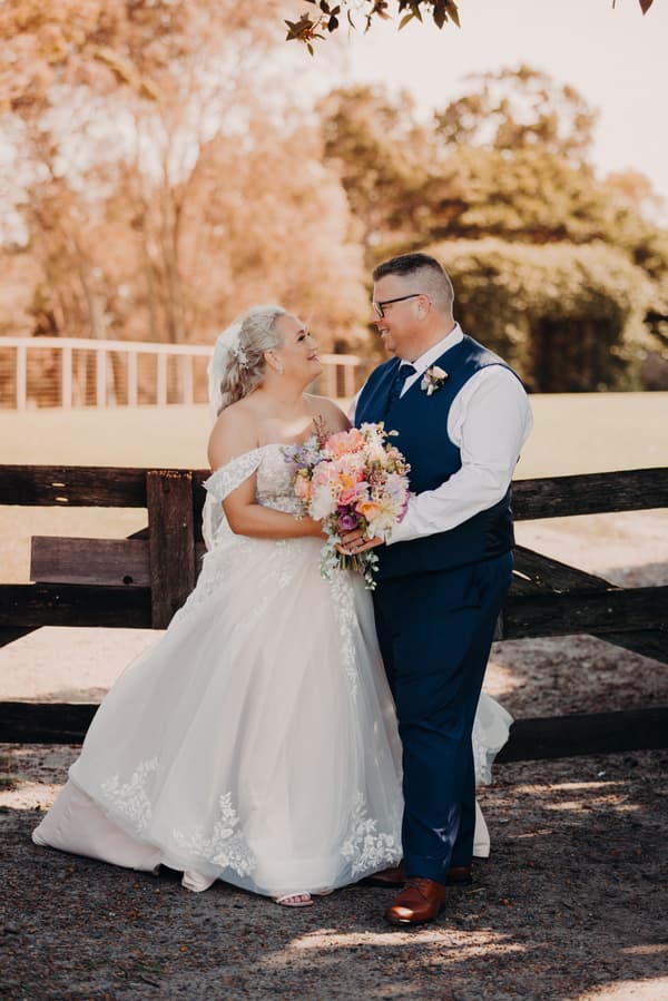 Bride Chantelle and groom Peter stand together holding a bouquet in front of a wooden fence at Sandstone Point Hotel, looking at each other and smiling.