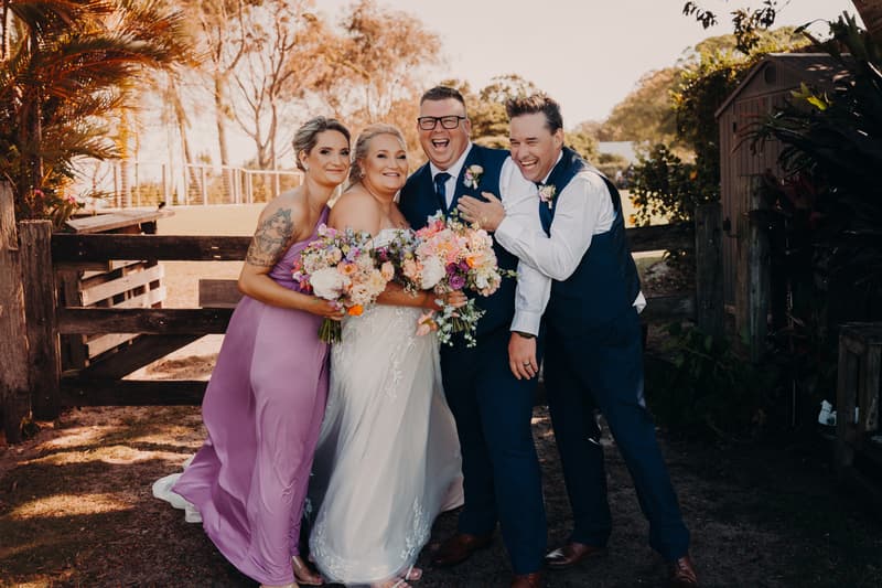 Chantelle the bride in a white wedding gown and Peter the groom in a navy suit pose with a bridesmaid in a lavender dress and a groomsman in a navy suit at Sandstone Point Hotel, all holding bouquets and smiling outdoors near a wooden fence.