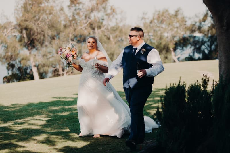 The bride Chantelle in a white wedding gown holding a bouquet and a drink, walking hand in hand with the groom Peter in a navy vest and tie, holding a bottle, outdoors on a grassy area at Sandstone Point Hotel.