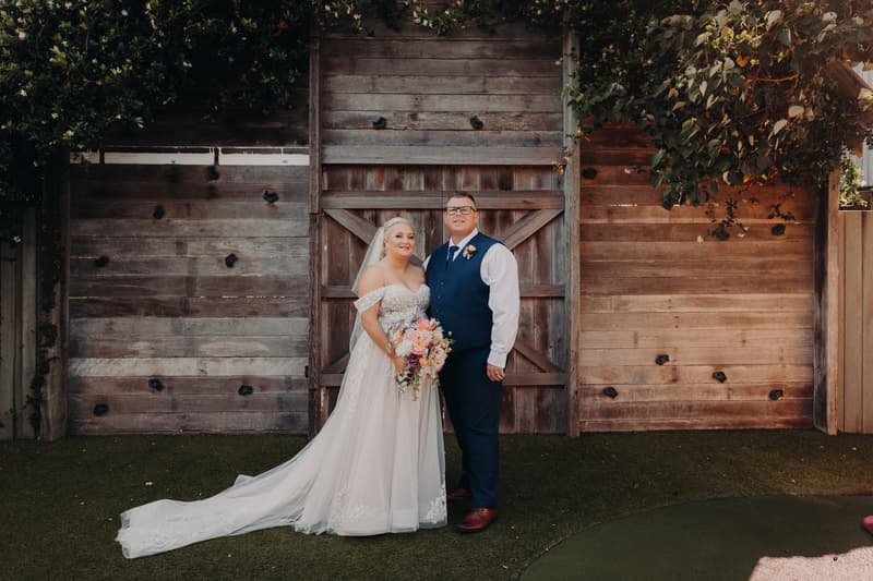 The bride Chantelle in a white wedding gown holding a bouquet and the groom Peter in a navy vest and white shirt stand together in front of a rustic wooden gate at Sandstone Point Hotel.