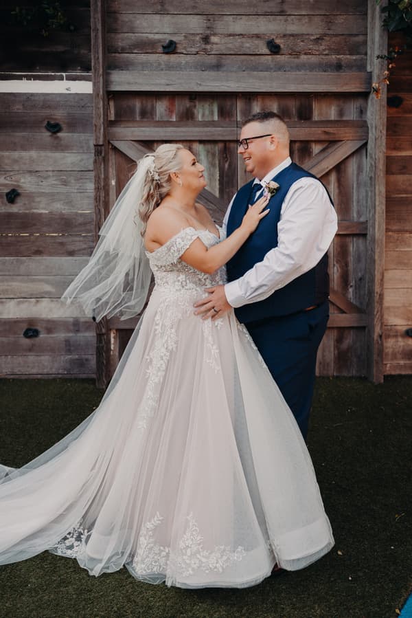 The bride Chantelle in a lace off-shoulder wedding gown and veil stands facing the groom Peter, who is wearing a white shirt, navy vest, and tie, in front of a rustic wooden door at Sandstone Point Hotel.