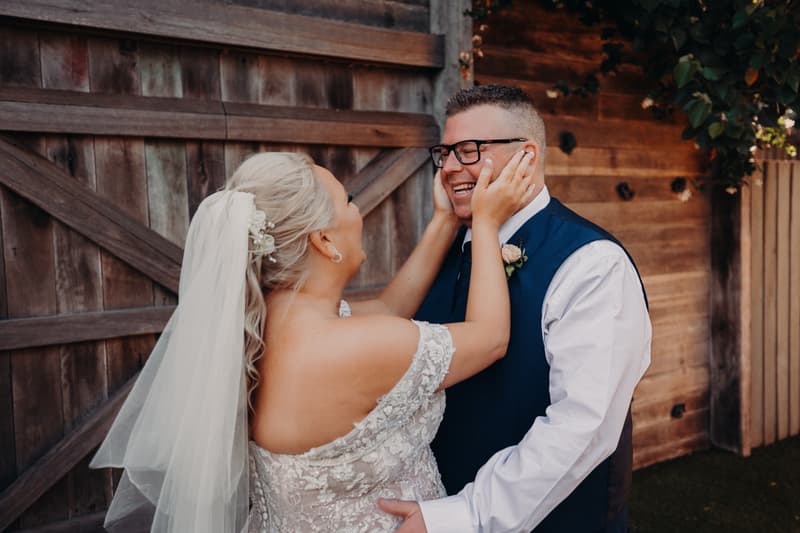 Bride Chantelle in a white lace off-shoulder gown and veil holds the face of groom Peter, who is wearing glasses, a white shirt, and a dark vest, posing together in front of a wooden fence at Sandstone Point Hotel.