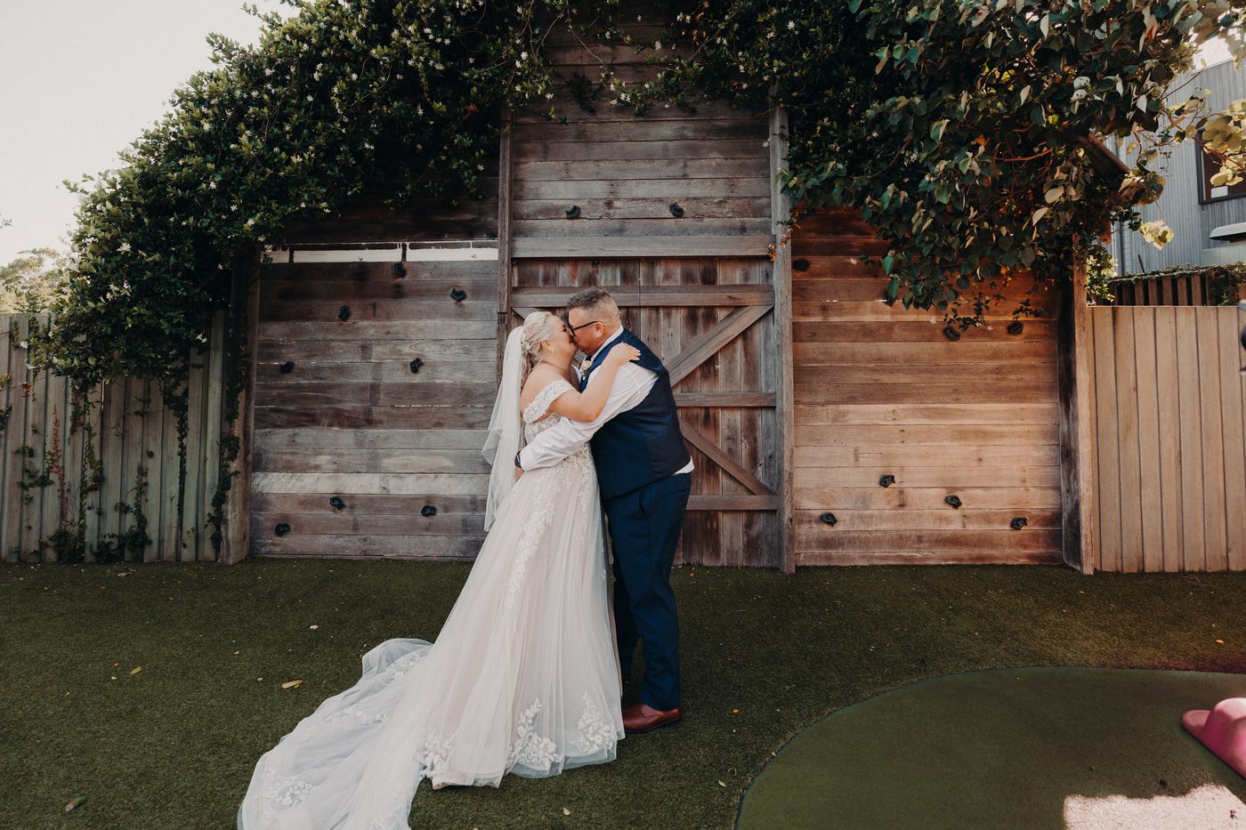 The bride Chantelle and groom Peter kiss in front of a rustic wooden gate. Wedding photography by She Said Yes.