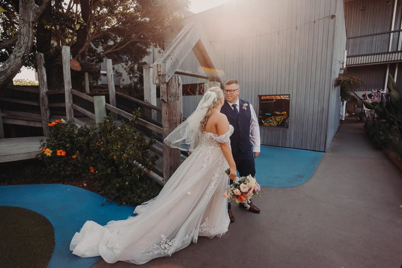 The bride Chantelle in a white lace wedding gown and veil holding a bouquet stands facing the groom Peter, who is wearing a navy vest and tie with a white shirt, outside near a wooden fence and a grey building at Sandstone Point Hotel.