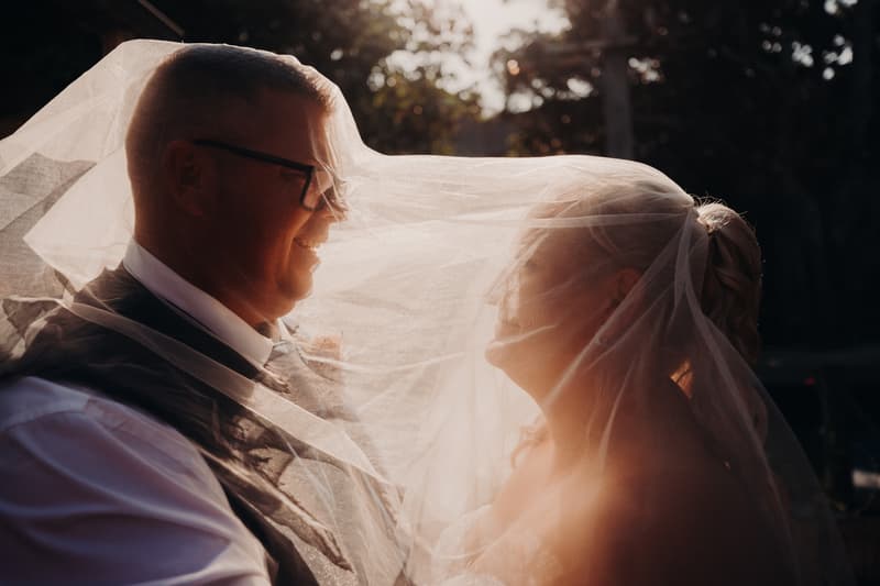 The bride Chantelle and groom Peter stand close together outdoors at Sandstone Point Hotel, with the bride's veil draped over both of them in soft sunlight.