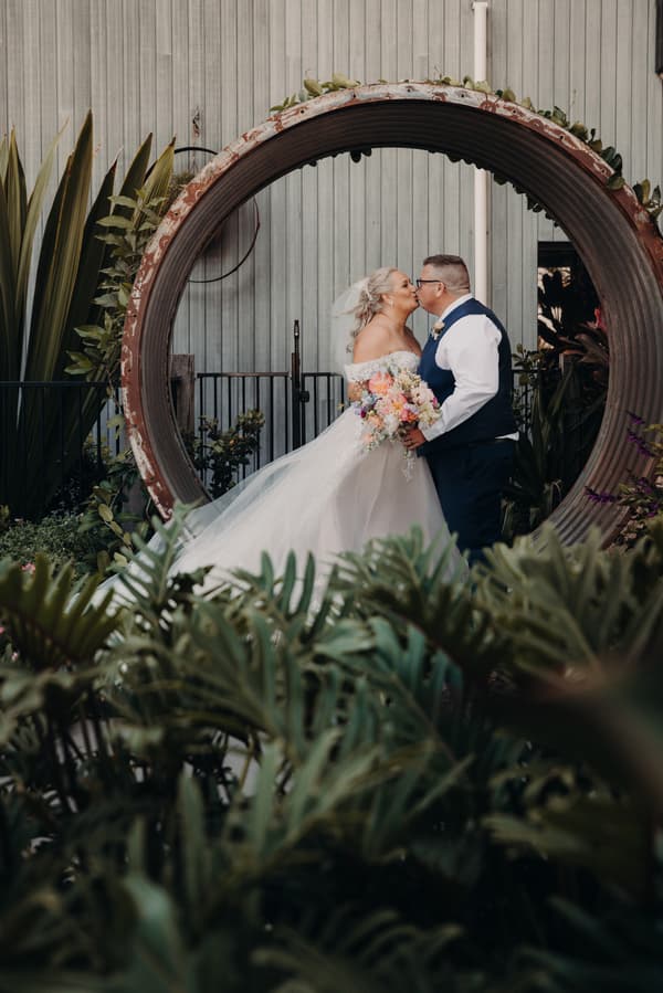 Bride Chantelle and groom Peter kiss while standing inside a large circular metal sculpture at Sandstone Point Hotel, surrounded by greenery. Chantelle wears an off-shoulder wedding gown and holds a colorful bouquet, while Peter wears a white shirt with a navy vest and pants.