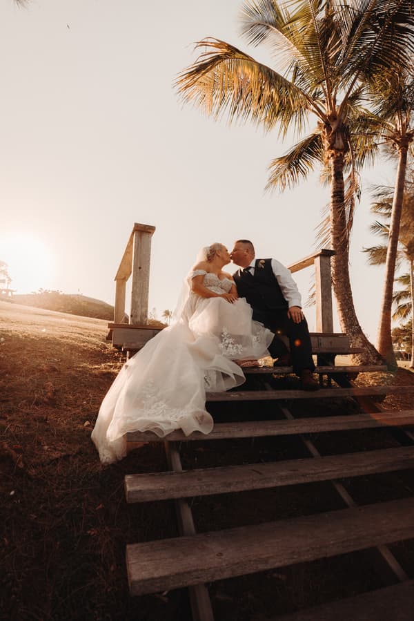 Bride Chantelle and groom Peter sit on wooden steps outdoors at Sandstone Point Hotel, sharing a kiss with palm trees and sunset in the background.