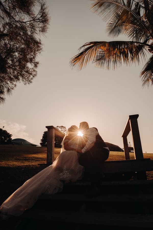 The bride Chantelle and groom Peter sit closely together on outdoor wooden steps at Sandstone Point Hotel during sunset, with the sun shining between their faces and palm trees overhead.