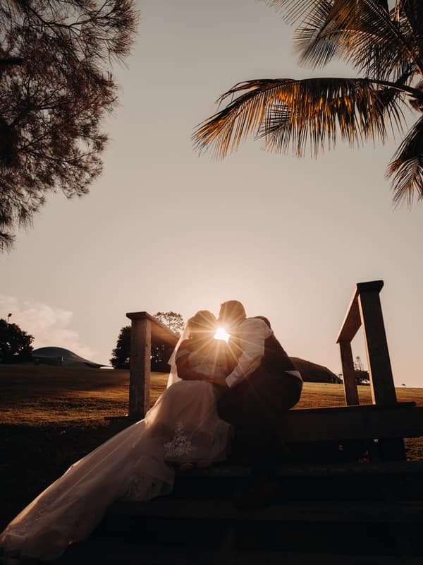 The bride Chantelle and groom Peter sit closely together on outdoor wooden steps at Sandstone Point Hotel during sunset, with the sun shining between their faces and palm trees overhead.