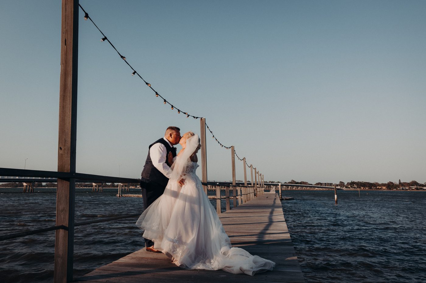 The bride Chantelle and groom Peter share a kiss on a wooden pier at Sandstone Point Hotel, with string lights overhead and water surrounding the pier under a clear sky.