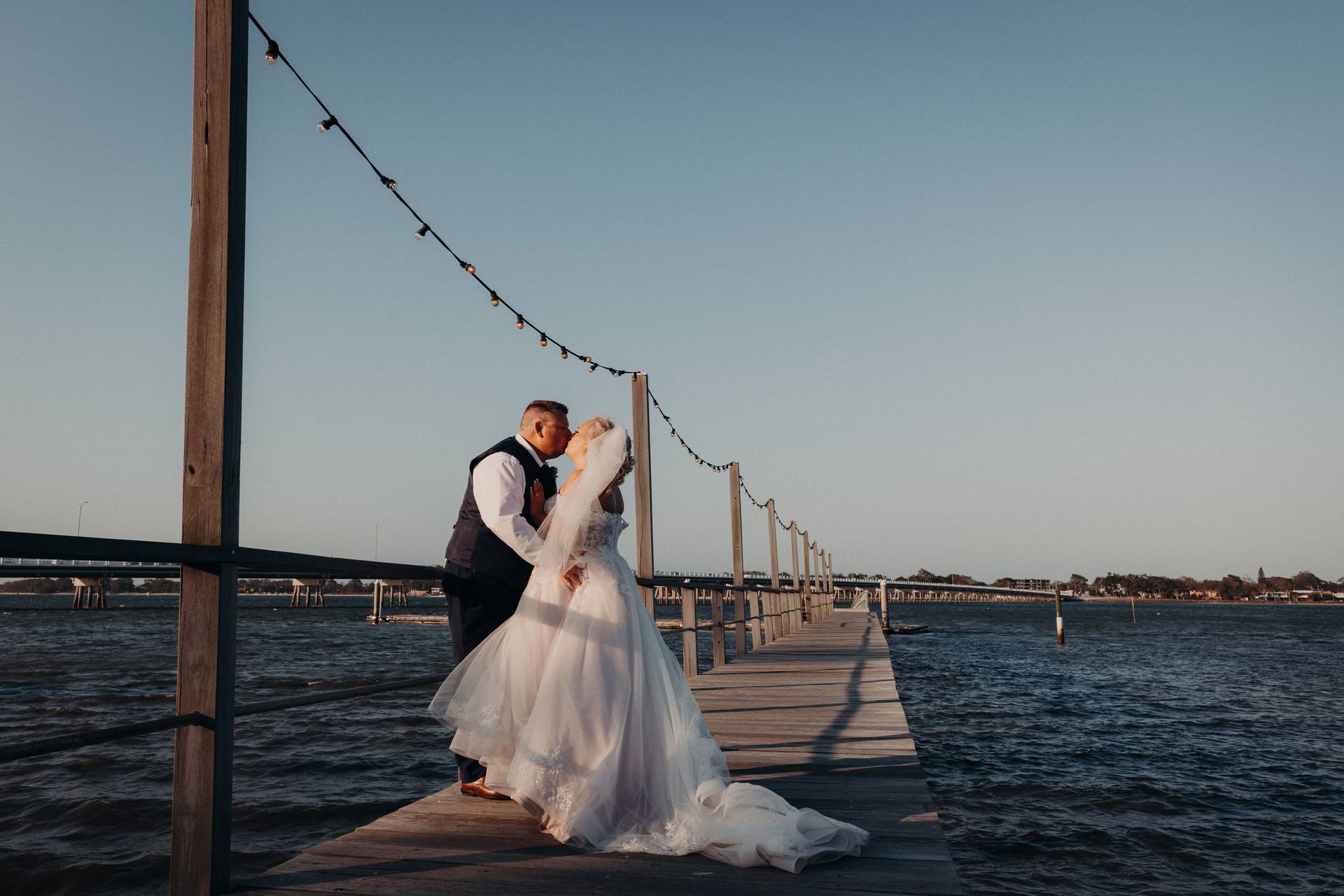 The bride Chantelle and groom Peter share a kiss on a wooden pier at Sandstone Point Hotel, with string lights overhead and water surrounding the pier under a clear sky.