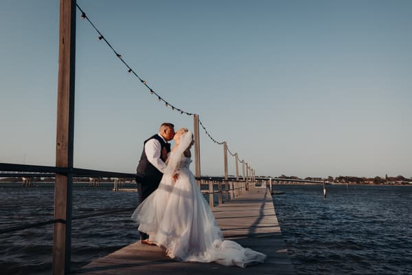 The bride Chantelle and groom Peter share a kiss on a wooden pier, with string lights overhead and water surrounding the pier under a clear sky. Elopement photographer Brisbane by She Said Yes.