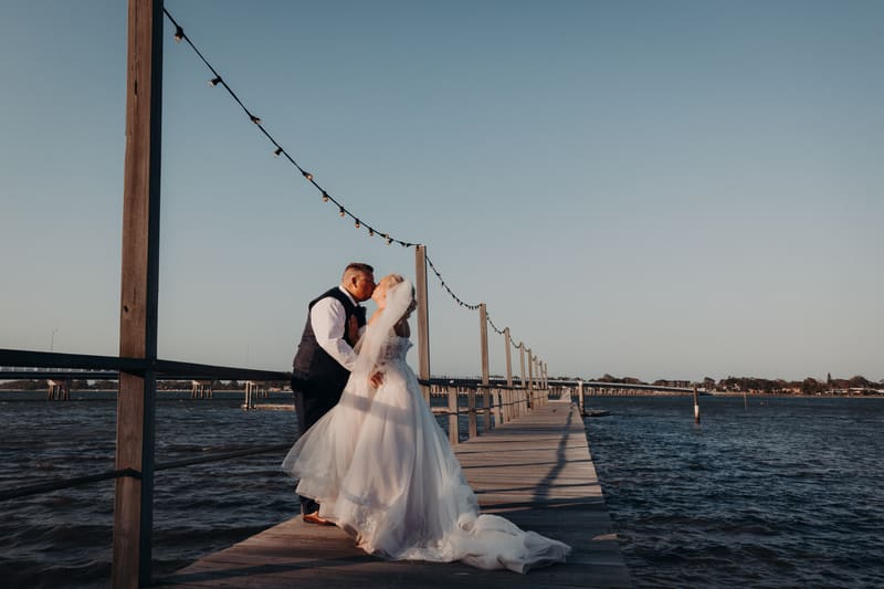 The bride Chantelle and groom Peter share a kiss on a wooden pier, with string lights overhead and water surrounding the pier under a clear sky. Destination wedding videographer by She Said Yes.