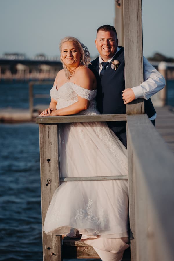 Chantelle the bride and Peter the groom pose together on a wooden railing overlooking water at Sandstone Point Hotel during their couple portraits session.