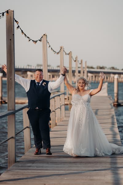 The bride Chantelle and groom Peter walk hand in hand on a wooden pier, both raising their free hands in celebration. Event videographer Brisbane by She Said Yes.
