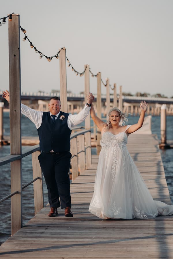 The bride Chantelle and groom Peter walk hand in hand on a wooden pier at Sandstone Point Hotel, both raising their free hands in celebration.