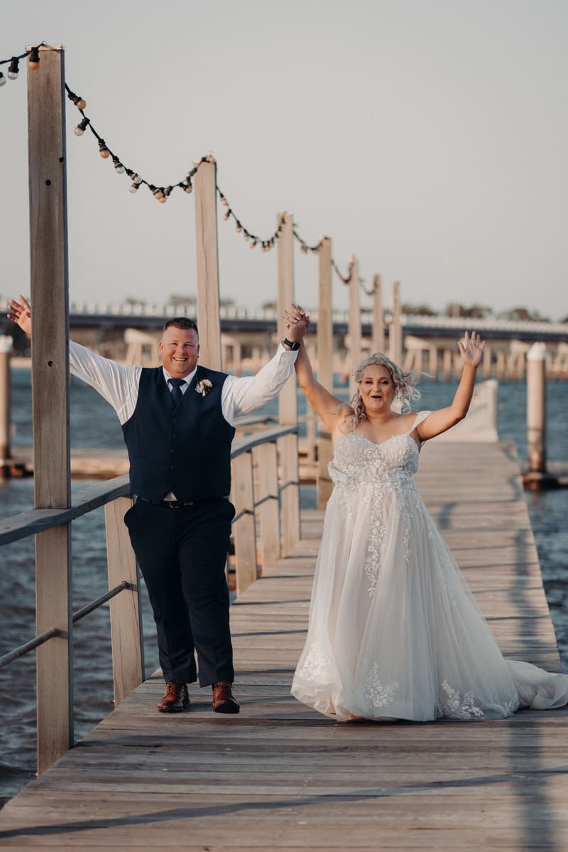 The bride Chantelle and groom Peter walk hand in hand on a wooden pier, both raising their free hands in celebration. Winter wedding light photography Brisbane by She Said Yes.