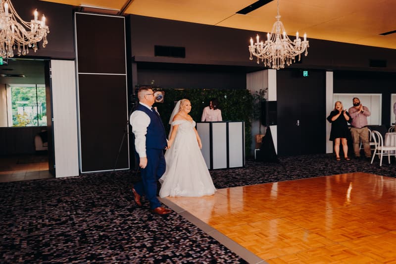 Bride Chantelle and groom Peter enter the reception room holding hands at Sandstone Point Hotel — Pumicestone Room, with guests clapping in the background.