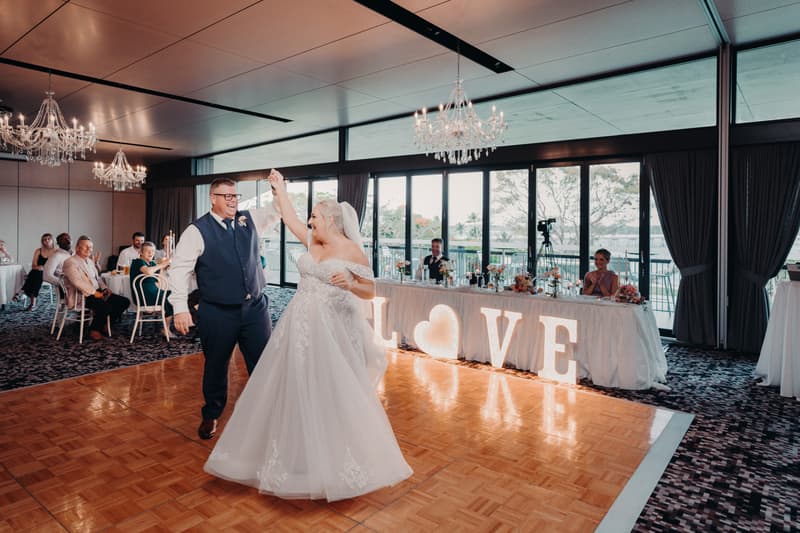 Bride Chantelle and groom Peter dance on the parquet dance floor at their wedding reception in the Pumicestone Room at Sandstone Point Hotel, with guests seated at tables and a head table decorated with illuminated LOVE letters behind them.