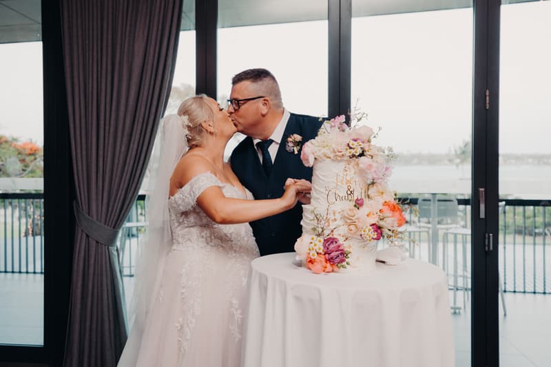 Bride Chantelle and groom Peter kiss beside their decorated wedding cake on a table at Sandstone Point Hotel — Pumicestone Room, with large windows and a balcony overlooking water in the background.